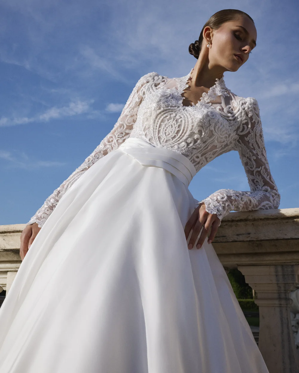 Sposa con corpetto di pizzo a maniche lunghe e gonna ampia, appoggiata a una balaustra sotto il cielo