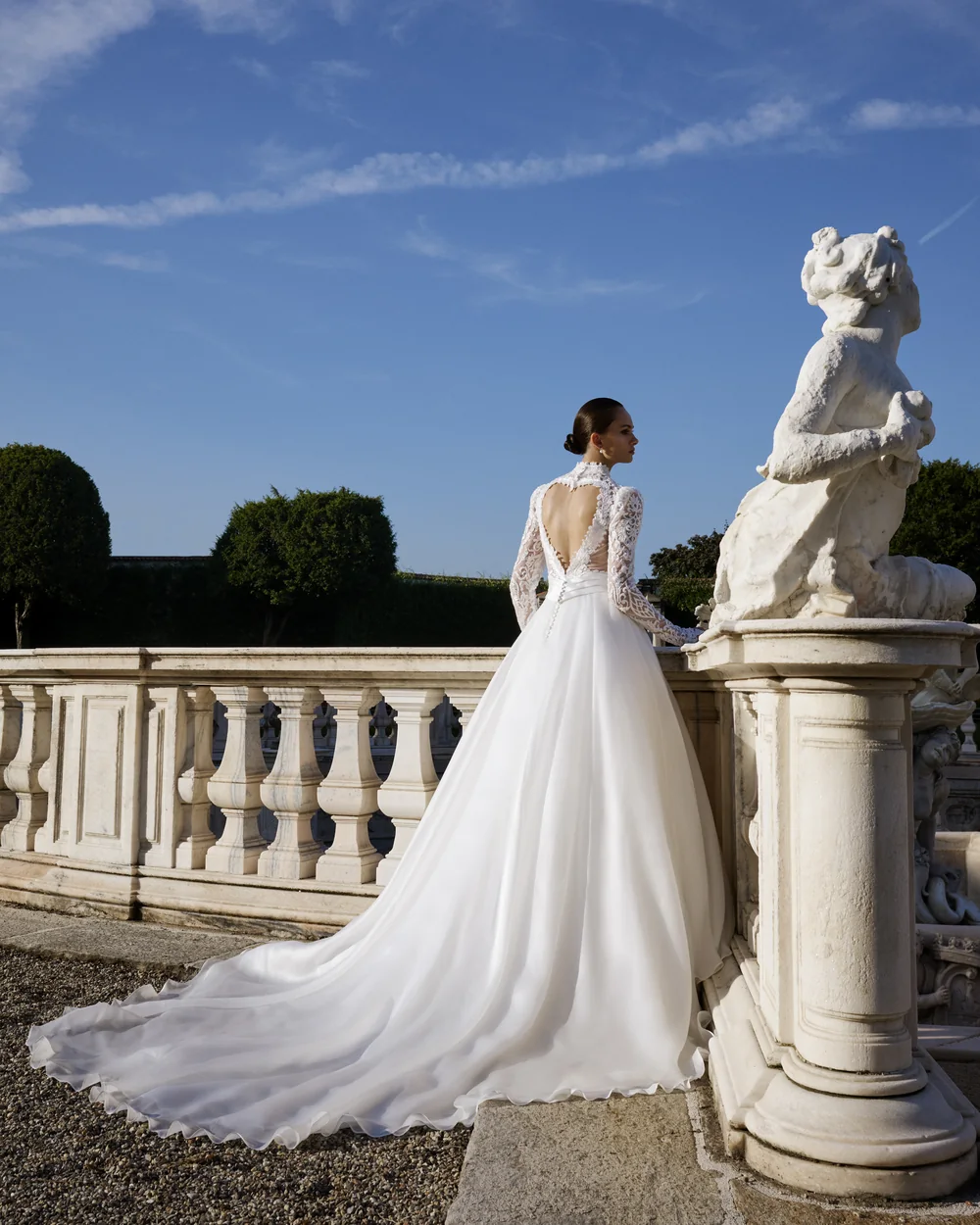 Sposa di spalle con corpetto di pizzo e lungo strascico, accanto a una statua su una balaustra