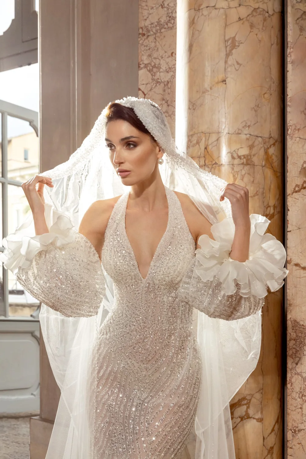 Sposa con il velo Molaro e abito ricamato di cristalli, accanto a una colonna di marmo rosa